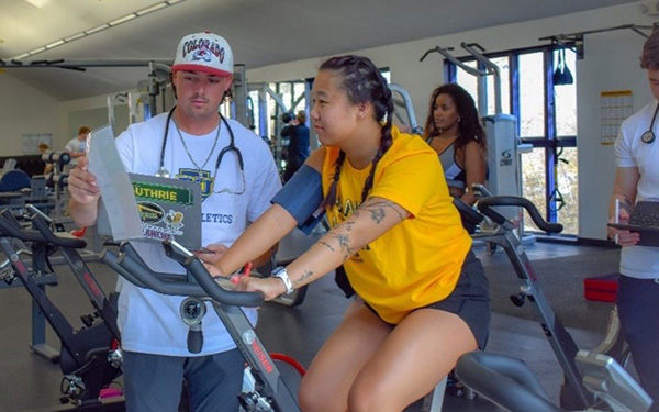 In the fitness center on the Northwest Denver campus, a student sits on an exercise bike with a blood pressure cuff on her arm while another student stands beside her wearing a stethoscope and shows her what is on his clipboard.