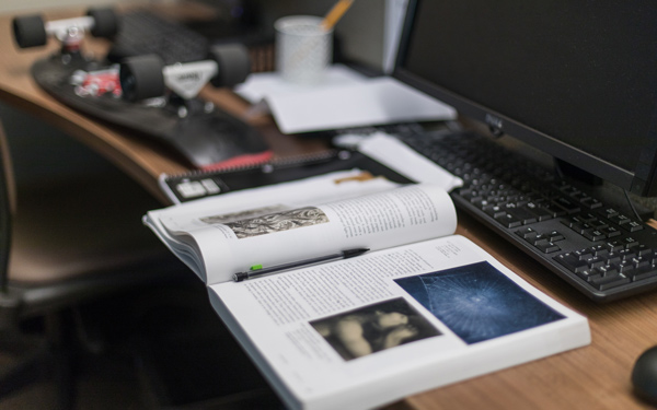 Texbook open on a desk with a computer keyboard and monitor. A skateboard can be seen in the background of the desk