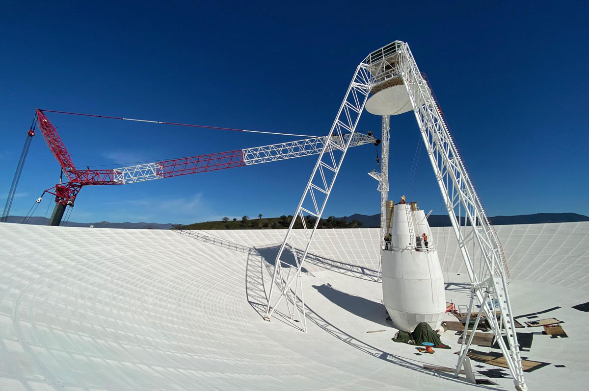 Deep Space Station 43 (DSS-43), a massive 70-meter-wide (230-foot) antenna at the Canberra Deep Space Network station in Australia, undergoing upgrades with workers and a large crane visible near the central cone housing transmitters and receivers. The towering structure, responsible for transmitting commands to NASA's Voyager 2 spacecraft, stands against a clear blue sky.