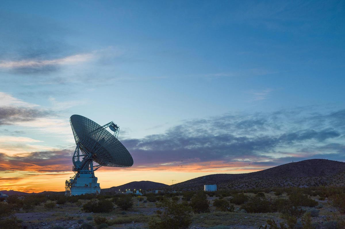 A large 70-meter-wide antenna known as Deep Space Station 14 (DSS-14) at the Goldstone Deep Space Communications Complex, silhouetted against a vibrant sunset with colorful skies and desert landscape in the background.