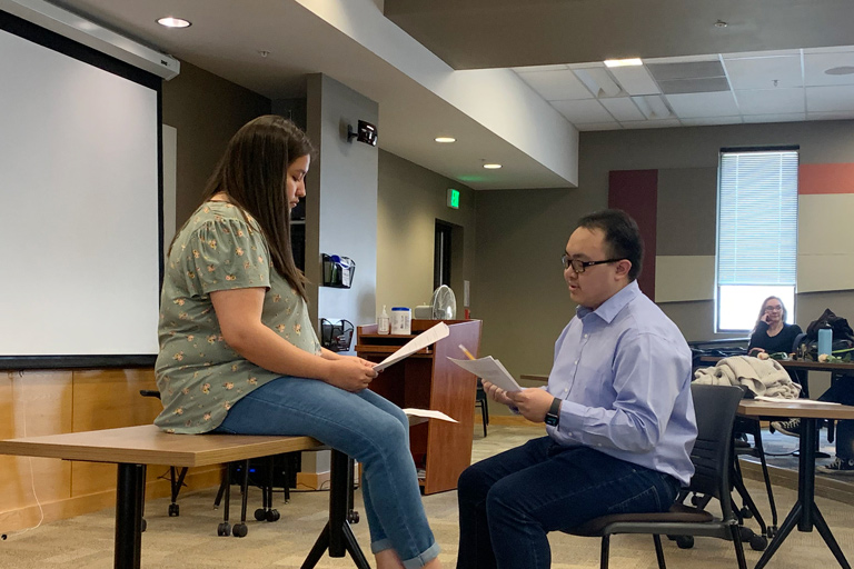 two students staging a play in front of their class, reading lines from their scripts