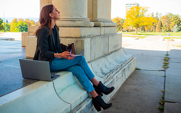 young professional woman writes on a notepad with her open laptop beside her while sitting in an urban park