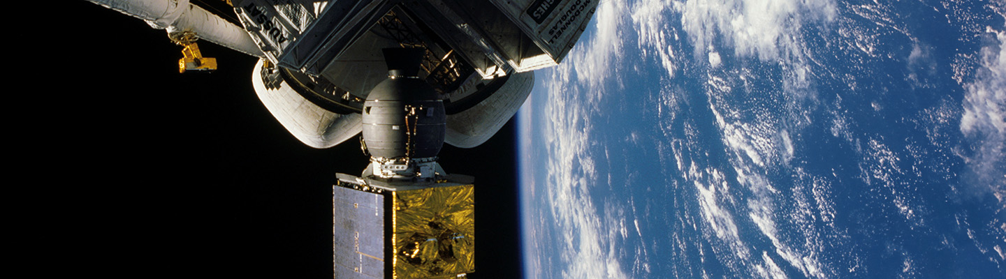 This image appears to be taken in space, showing part of what looks like a satellite or spacecraft component. In the foreground, there's a mechanical structure with a metallic grey body and what appears to be a gold-colored satellite or payload attached to it. The background shows a stunning view of Earth from space, with the characteristic curvature of the planet visible against the black of space. You can see the blue oceans and white cloud patterns over the Earth's surface. The image has very high quality and clarity, suggesting it was likely taken during a NASA or other space agency mission.