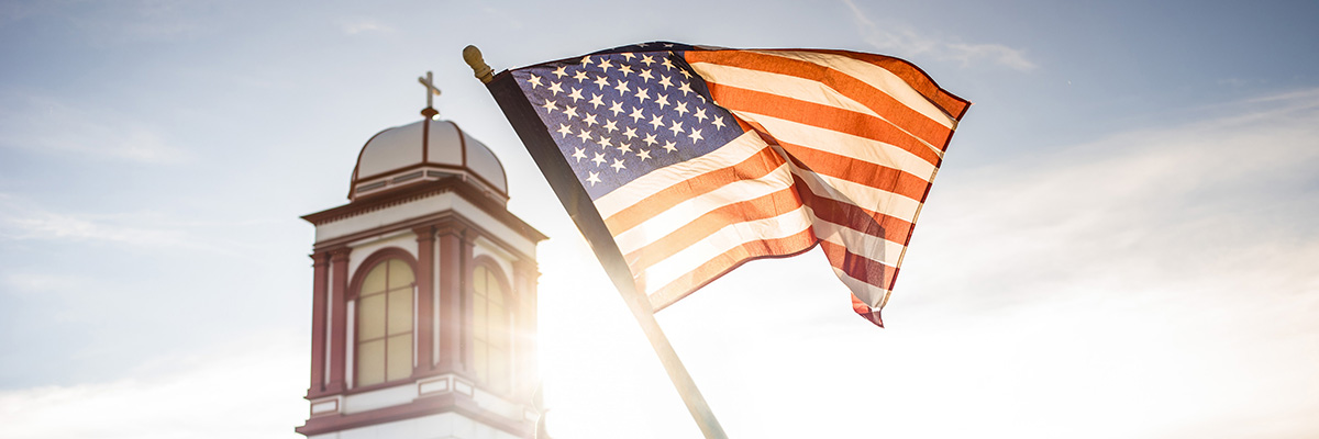 A backlit American flag waves in the wind next to Regis University bell tower topped with a cross. The sun creates a bright glow behind the bell tower. The flag's stars and stripes are clearly visible against a bright, slightly cloudy sky, creating a patriotic and spiritual composition.