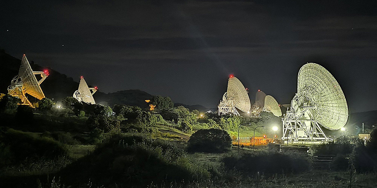 A nighttime photograph of multiple large radio telescope dishes arranged along a hillside. The large parabolic antennas appear illuminated in white/yellow light against the dark sky, with red aircraft warning lights visible on their tops. The telescopes are surrounded by vegetation and appear to be part of a radio astronomy facility or deep space communications complex.