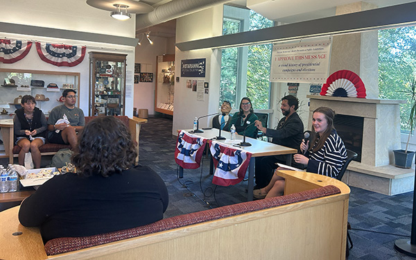 A panel discussion in a well-lit room decorated with patriotic-themed banners and memorabilia. Four panelists sit at a table with microphones, engaging with the audience. 