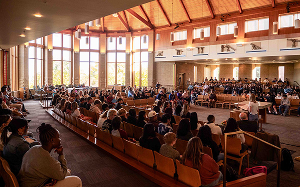 A large gathering in a spacious auditorium with high wooden ceilings and tall windows allowing natural light to fill the room.