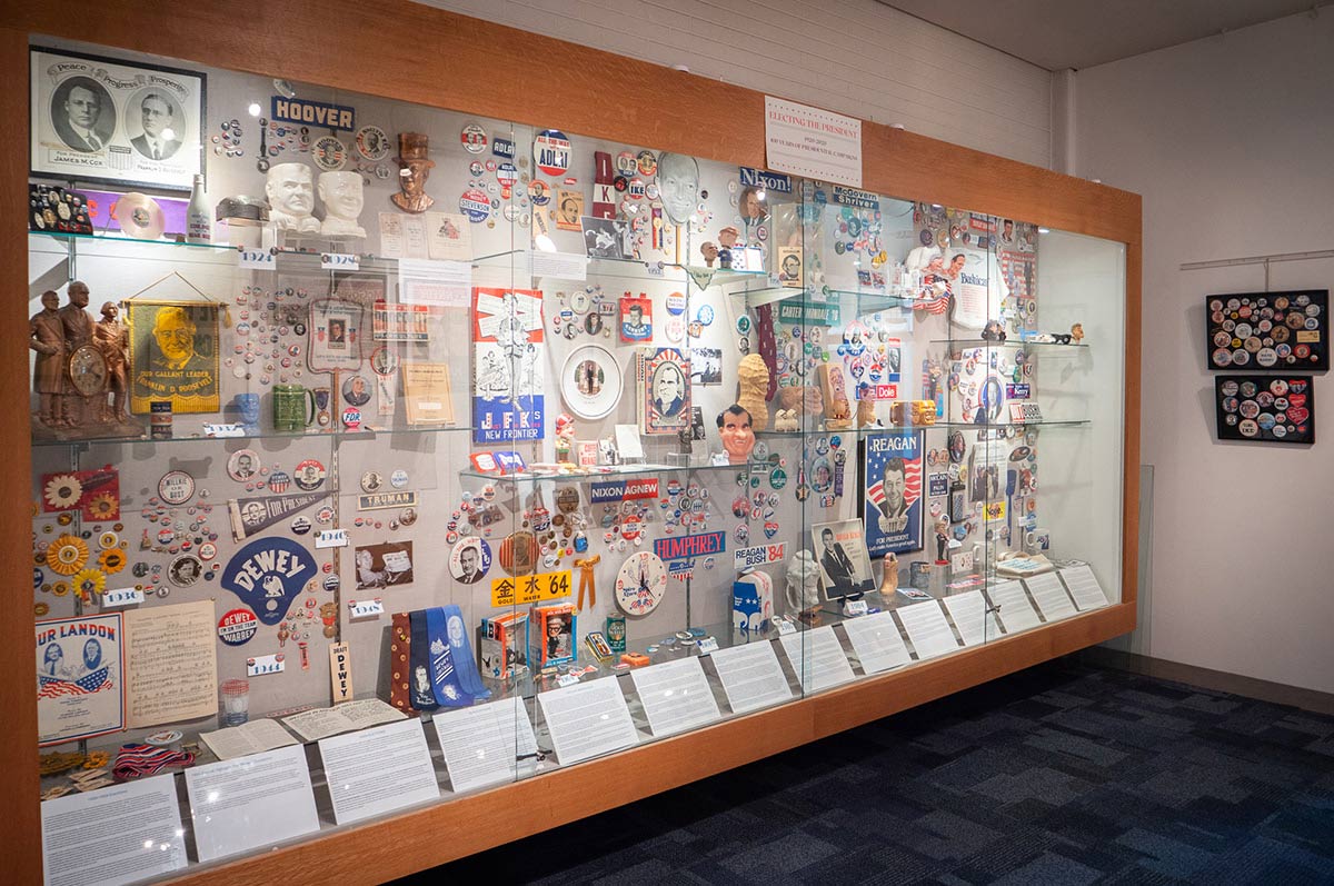 A large museum display case featuring an extensive collection of American presidential campaign memorabilia spanning several decades. The display includes campaign buttons, posters, photographs, banners, and other political ephemera from various presidential candidates and elections.