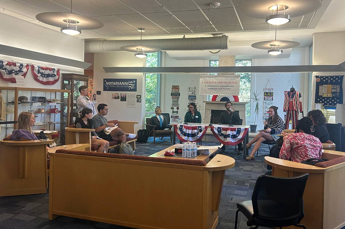 A group of people gathered in a room with American patriotic decorations, including red, white and blue bunting. Several speakers, appearing to be election officials, are seated at the front facing an audience seated on curved benches. There are display cases visible along the walls containing political memorabilia. 