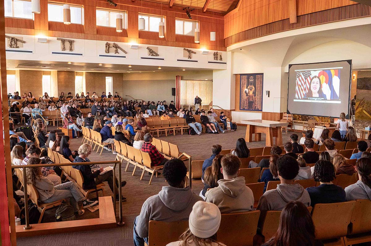 A chapel filled with students watching a video presentation. The room features wooden panels, high ceilings with pendant lights, and decorative friezes along the walls. Students are seated in rows of wooden chairs, facing a projection screen showing a speaker.