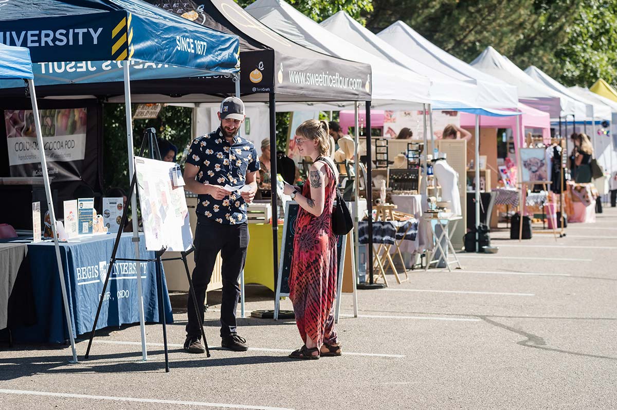 a shopper talks to a vendor beside a row of tents where artisans sell their goods and the Horseshoe market in a parking lot on the regis campus.