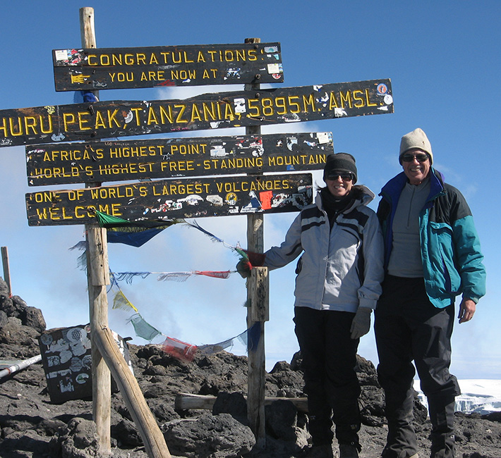 Two people stand in front of a sign on a mountain