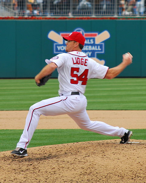 Lidge, Philadelphia Phillies pitcher, wearing a red and white uniform with #54, captured mid-motion on the pitcher's mound. He is in full stride with his arm reared back, preparing to throw a pitch