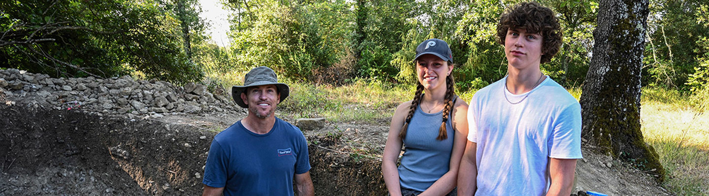 Lidge standing in a dug-out excavation site, smiling at the camera, wearing a sun hat and t-shirt; alongside his daughter, who has braided hair, a ball cap, and a tank top; and his son, dressed in a white t-shirt and chain necklace