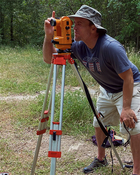 Lidge appears focused looking through a device used to measure coordinates and elevation, in a wooded grassy area, wearing a sun hat, shorts, T-shirt, and hiking boots