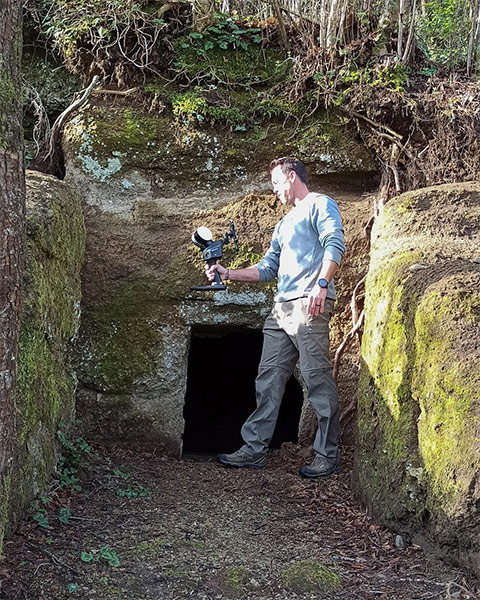 Lidge standing in front of an ancient Etruscan tomb, surrounded by mossy rocks, tree roots, and ground covered in mulch and vines. He is dressed in hiking boots and outdoor apparel, holding a hand-held Lidar device extended in front of him, appearing focused on scanning the site