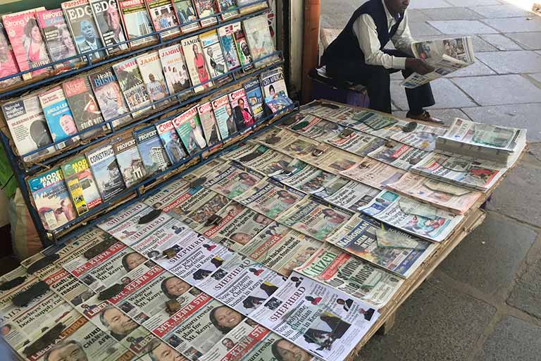 Table full of magazine in the middle. Behind it is a stand with more magazines. A person sit nexts to those as they ready one of the papers