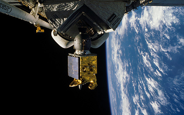 This image appears to be taken in space, showing part of what looks like a satellite or spacecraft component. In the foreground, there's a mechanical structure with a metallic grey body and what appears to be a gold-colored satellite or payload attached to it. The background shows a stunning view of Earth from space, with the characteristic curvature of the planet visible against the black of space. You can see the blue oceans and white cloud patterns over the Earth's surface. The image has very high quality and clarity, suggesting it was likely taken during a NASA or other space agency mission.