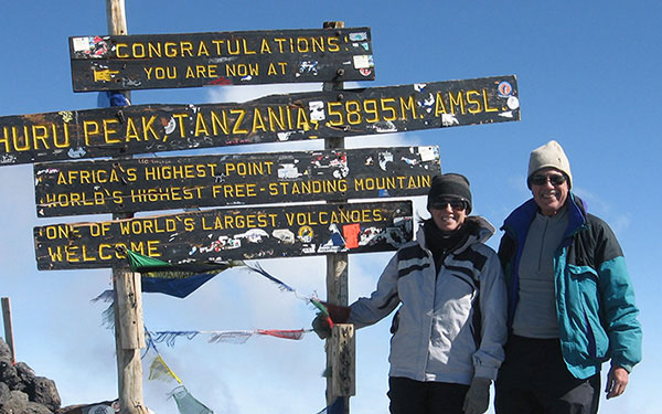 Two people stand in front of a sign on top of a mountain