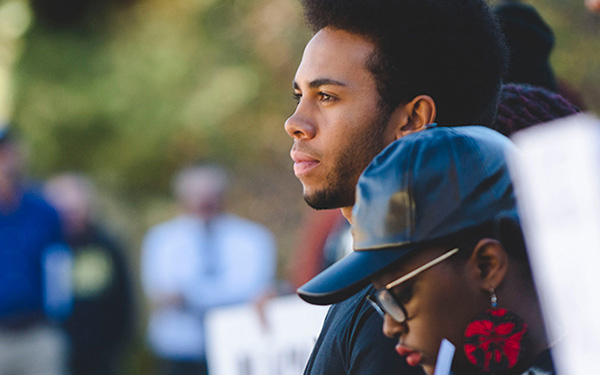 A man and woman attending a civil rights event; the man gazing into the distance, and the woman, wearing glasses and a hat, looking downward; both appear solemn and in solidarity; other attendees are visible in the background