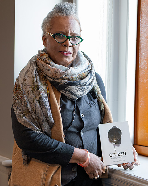 Radkine, leaning against a window sill with sunlight coming through, holding her book, Citizen: An American Lyric