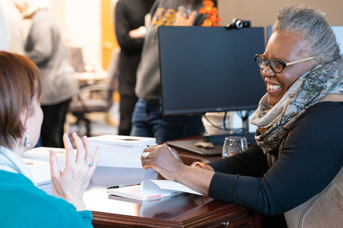 Radkine seated at a desk with a copy of her book open in front of her and a Sharpie marker beside it, ready to sign. She is smiling and looking at another woman seated nearby, who is speaking with animated hand gestures