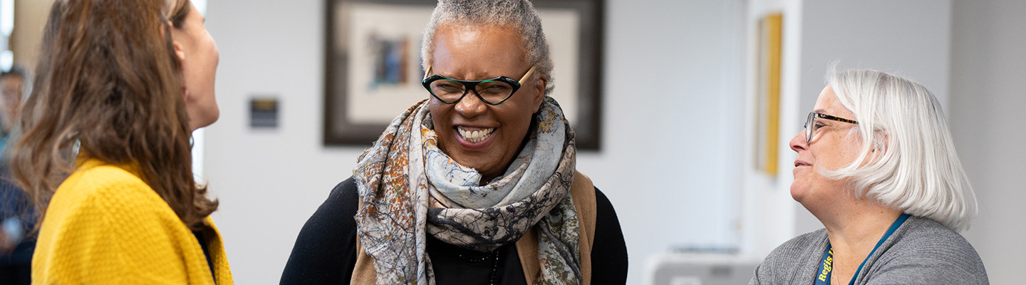 Radkine, wearing glasses and scarf, standing with two other women, having conversation and smiling