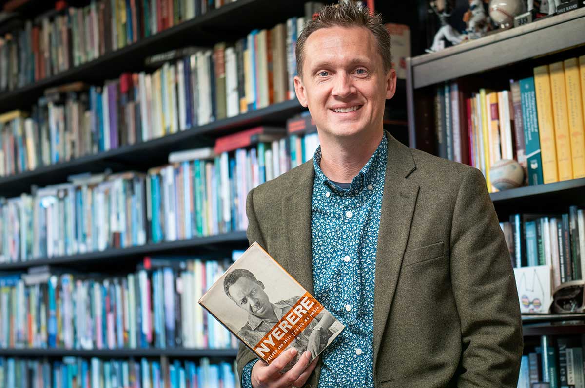 Ethan Sanders stands in front of a large, full bookshelf while holding a book titled "Nyerere" and featuring a photo of Julius Nyerere, Tanzania's first president