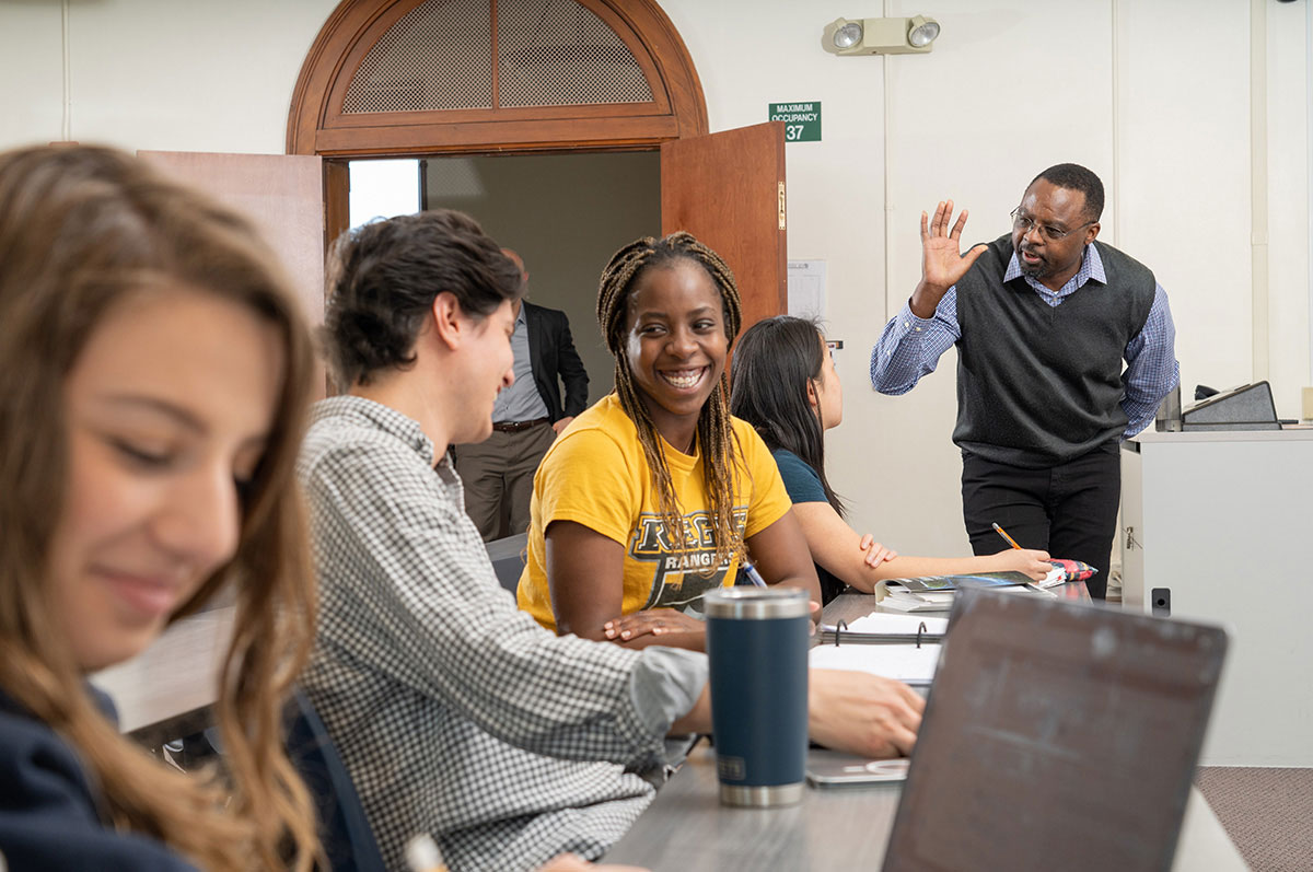 Kinoti talks animatedly to a student in a classroom while other students work in pairs on a class project