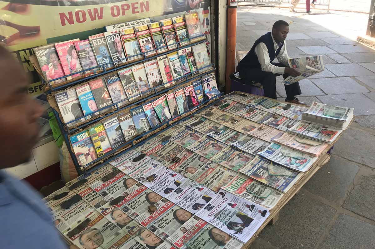 A man sits reading a newspaper beside a news stand on the sidewalk in Myanmar