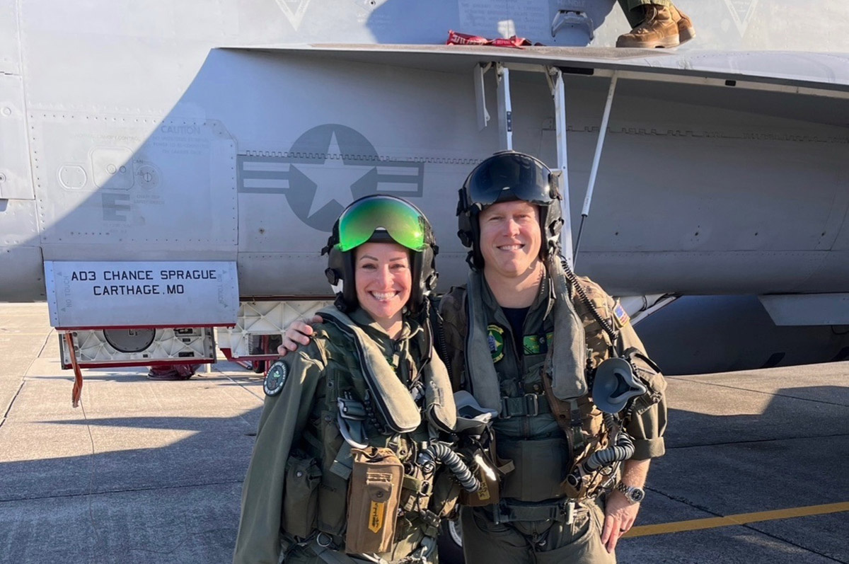Daphne Ryan and a Navy pilot wear flight gear and stand side by side on the tarmac in front of a Navy plane. Writing on the plane reads "AD3 Chance Sprague Carthage, MO"