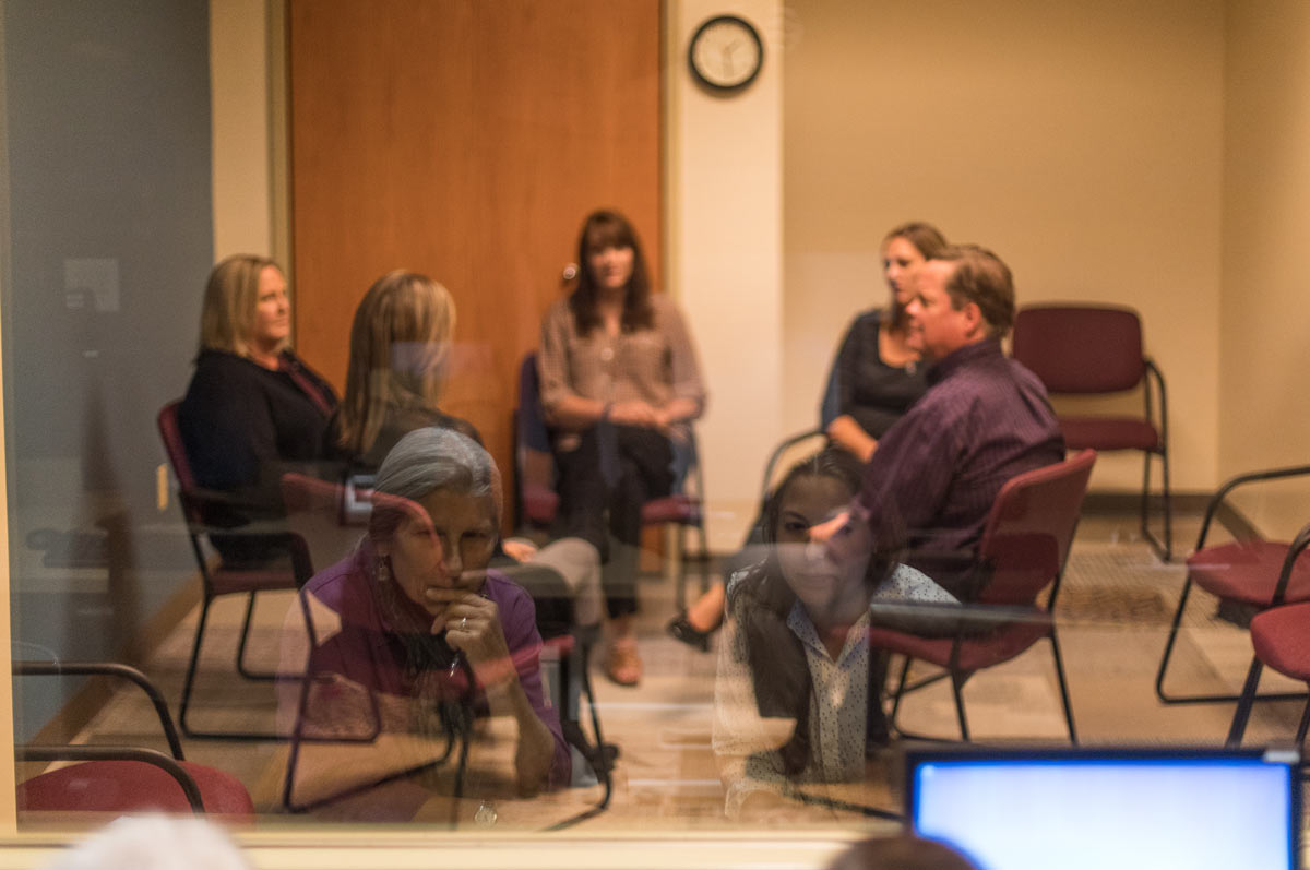 Five people are seated behind glass in a group therapy session. Reflected in the glass are a Regis student and Regis counseling professor observing the therapy session.