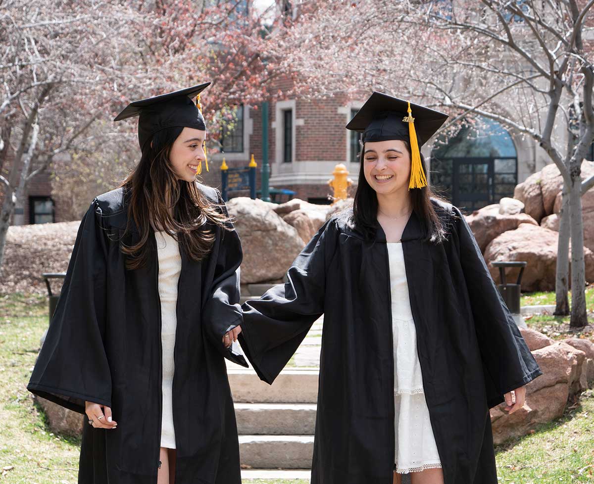 Isabella and Katelyn De Leon wear their graduation regalia while holding hands and smiling on the quad on the Northwest Denver campus