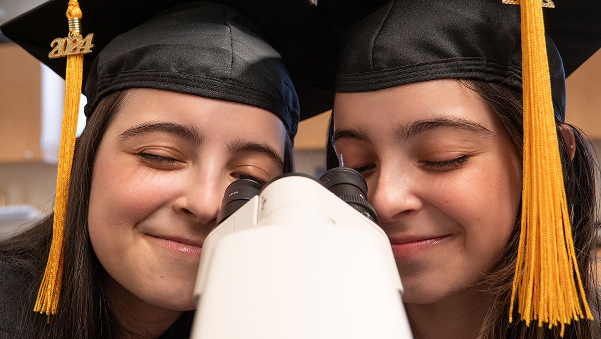 Isabella and Katelyn De Leon looks through a mircroscope while wearing their 2024 graduation caps in a biology lab on the northwest denver campus