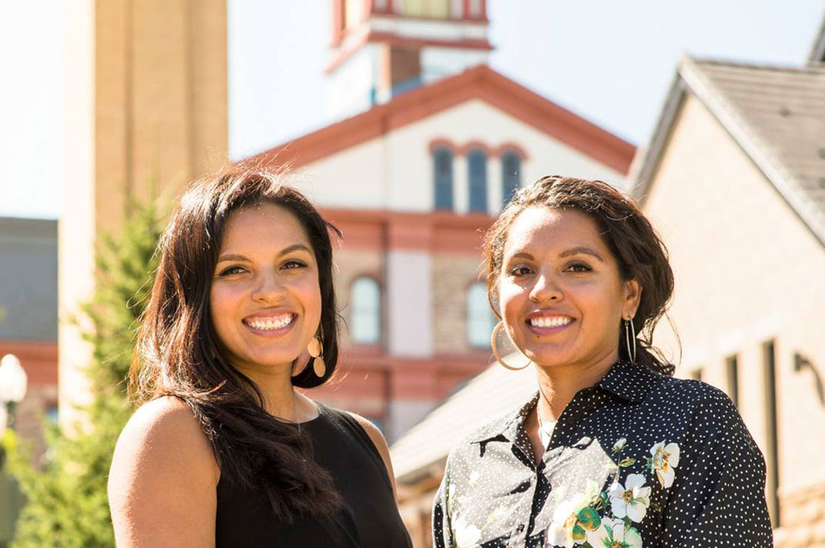 Diana and Denise Lopez stand side-by-side, smiling on the Regis Northwest Denver Campus with Main Hall and the Chapel in the background