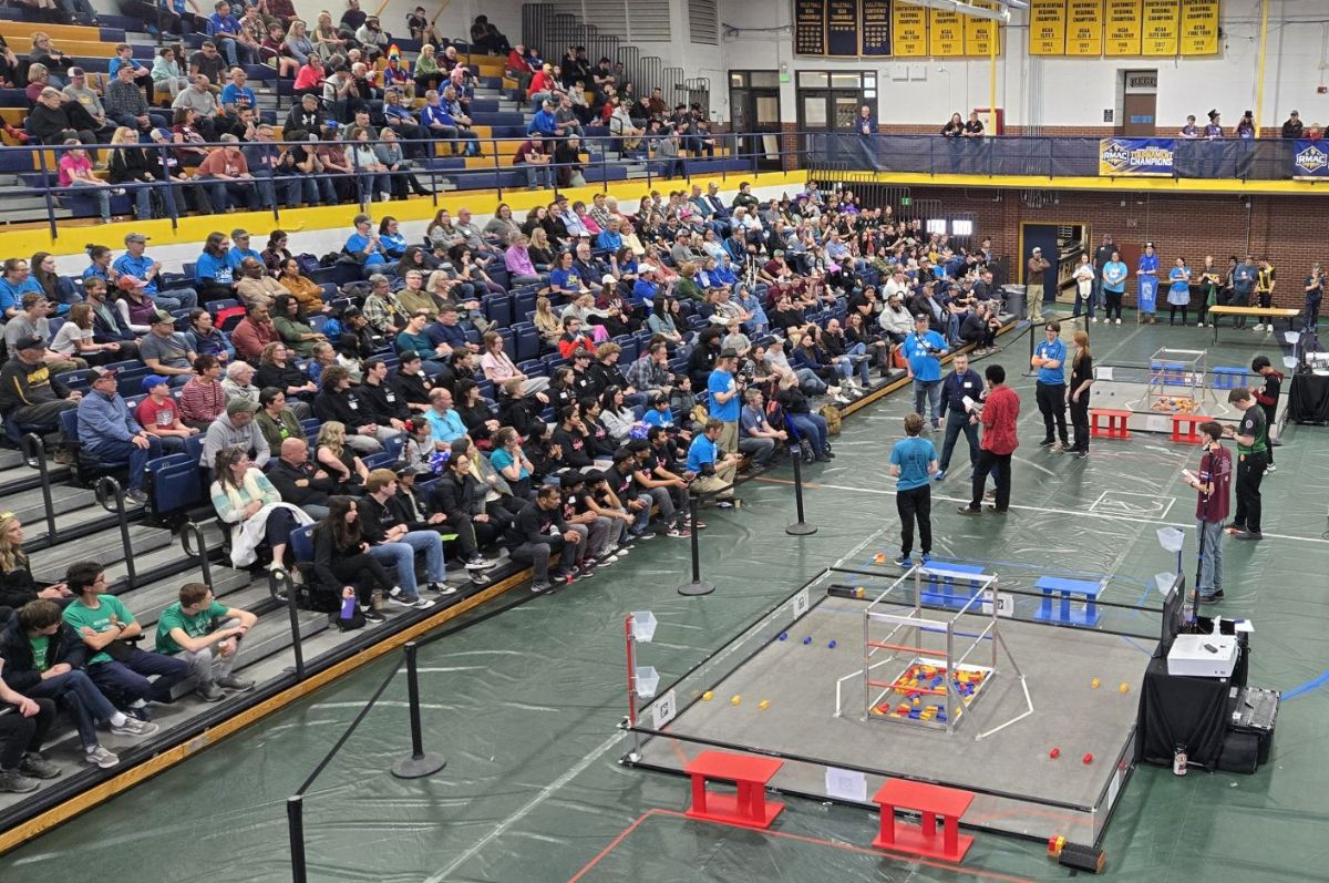 a packed gymnasium hosting a robotics competition, with bleachers full of spectators watching teams in matching shirts compete using robots on a playing field