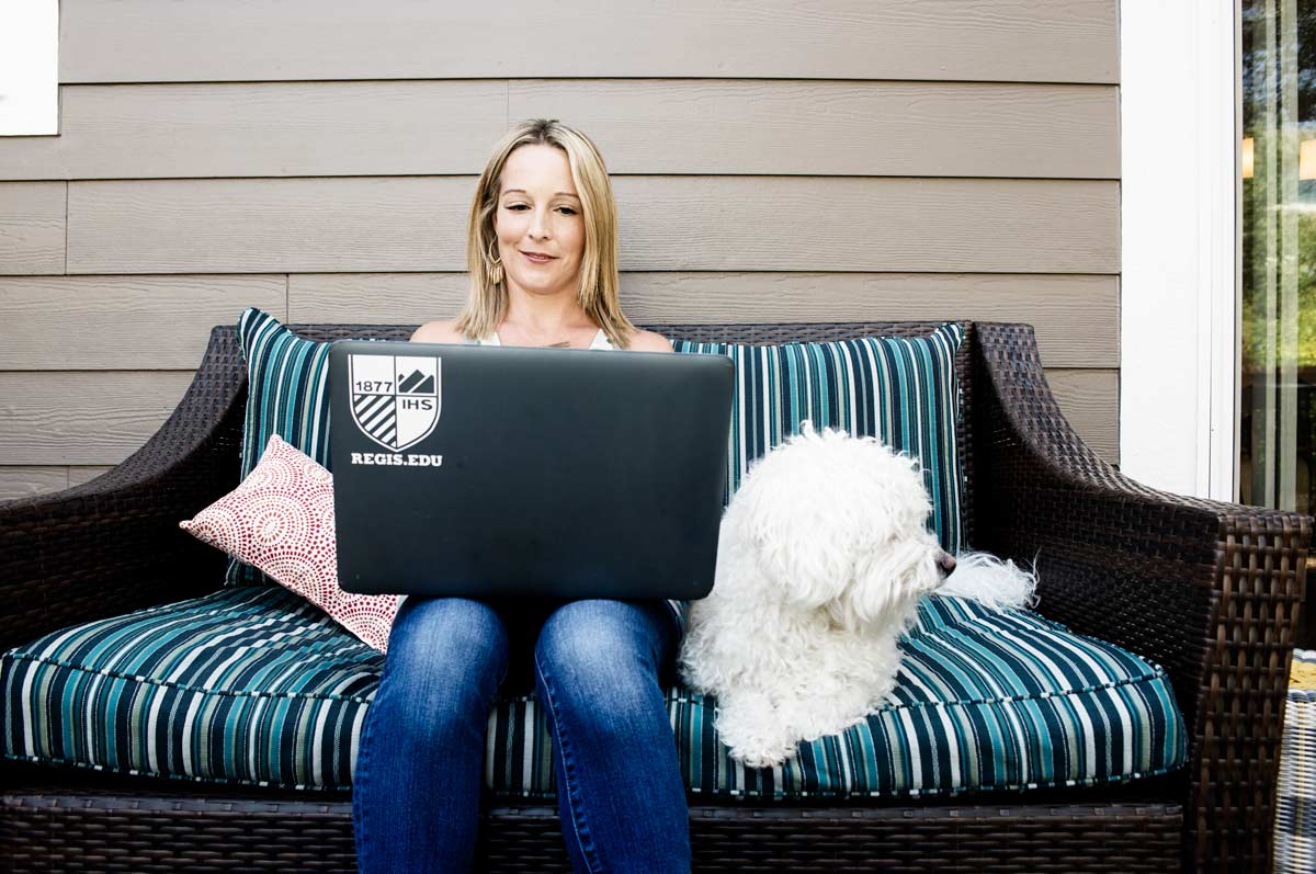 A student sits next to a dog on a patio sofa with a laptop with a Regis University sticker open on her lap.