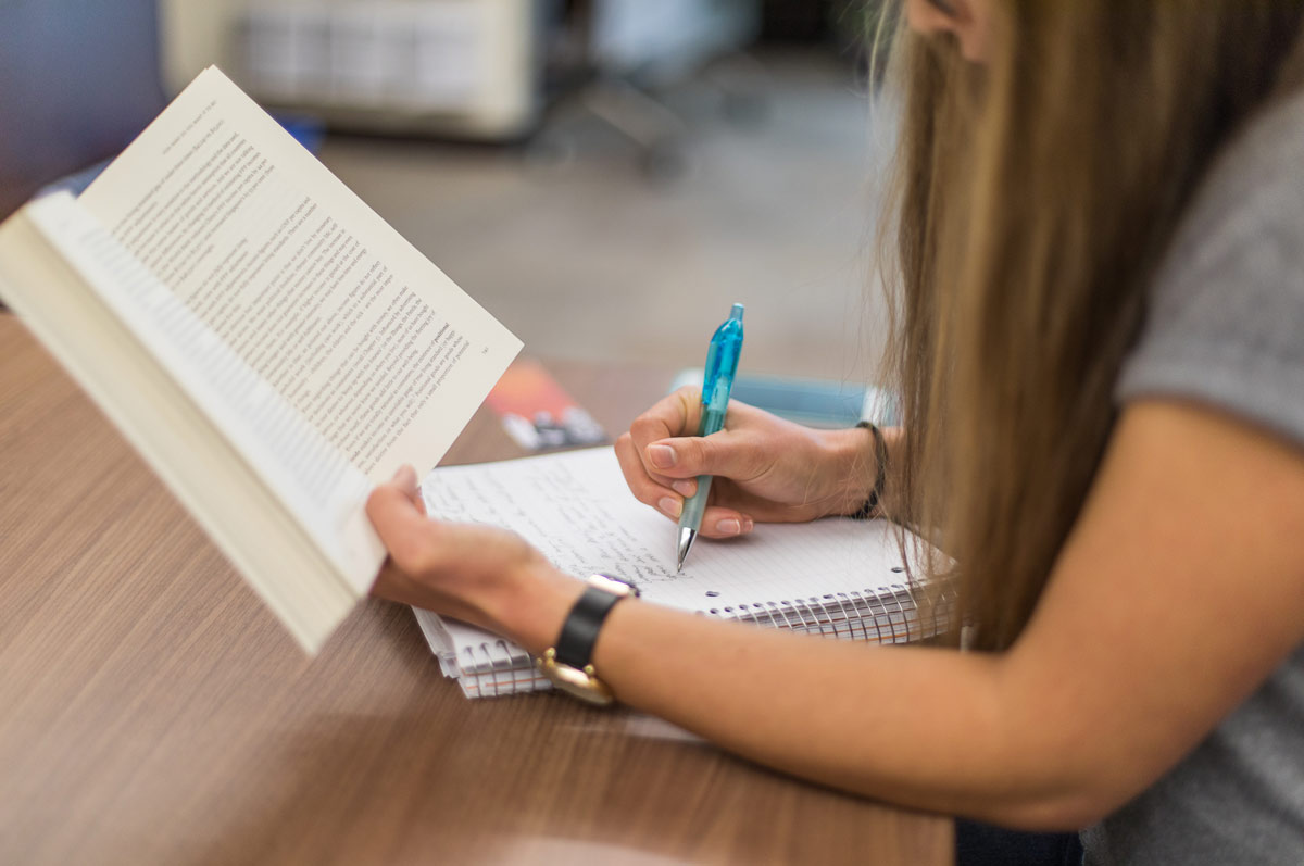 A student is seated in the Regis library holding a book in their left hand and taking notes in a notebook with their right hand.